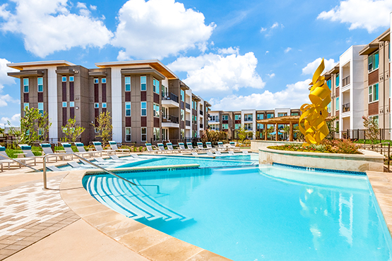 Pool with lounge chairs. A tall yellow sculpture in the flower bed. Apartment buildings in the background.